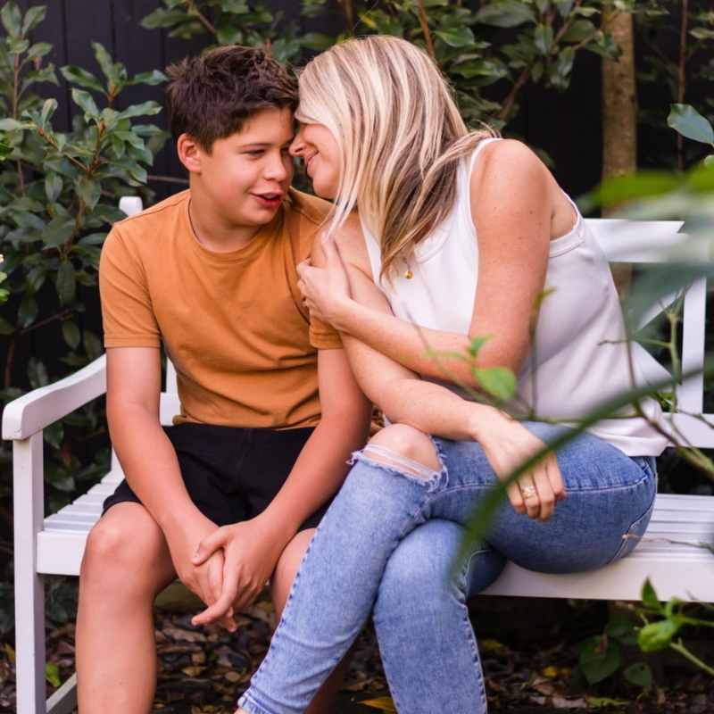 A boy and his mum are sitting on a white outdoor bench among green plants, leaning in and smiling.
