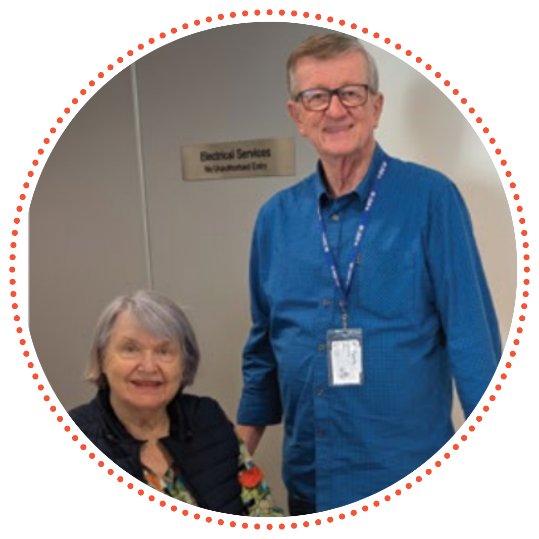 A photo of Greg and Phyllis on a shift at their local Lifeline Queensland Crisis Support Centre. They're smiling gently with Phyllis sitting in a chair and Greg standing comfortable beside her.