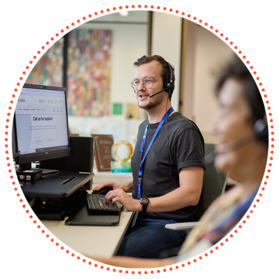 A Crisis Supporter Volunteer wearing a headset and glasses works at a computer, with another Crisis Supporter in the foreground also wearing a headset.